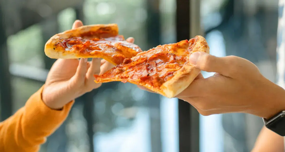 Dwie ręce podają sobie kawałki pizzy. Czy pizza jest zdrowa? Zależy od dodatków i sposobu przygotowania.