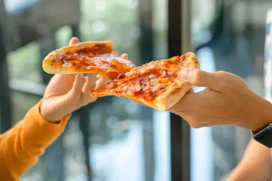 Dwie ręce podają sobie kawałki pizzy. Czy pizza jest zdrowa? Zależy od dodatków i sposobu przygotowania.