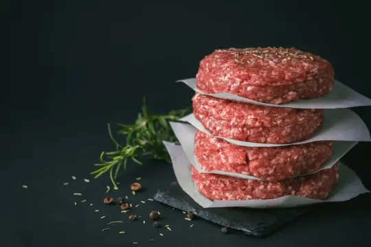 Stos świeżego mięsa na burgery, przyprawionego ziołami, obok gałązki rozmarynu i pieprzu.