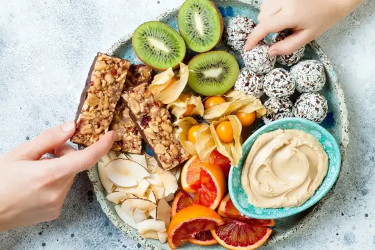 Talerz pełen zdrowych przekąsek: batony musli, kulki kokosowe, kiwi, physalis, pomarańcze i pasta orzechowa.